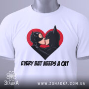 Футболка бетмен кішка кохання, біла, складена на столі, фон світло-сірий, принт з червоним серцем, на якому поцілуються персонажі Бетмен і Кішка, з етикеткою зеленого кольору.