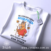 Футболка без подарунків зрада. Білий колір, зображена зверху на столі, фон світло-сірий. Принт з мультяшним конем у червоному халаті, що тримає палицю, з пташками навколо, на етикетці зелена стрічка.