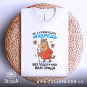 Футболка без подарунків зрада. Білий колір, зображена зверху на плетеному кошику, фон світло-сірий. Принт з мультяшним конем у червоному халаті, що тримає палицю, з пташками навколо.