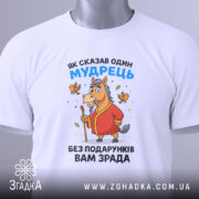 Футболка без подарунків зрада. Білий колір, зображена зверху на столі, фон світло-сірий. Принт з мультяшним конем у червоному халаті, що тримає палицю, з пташками навколо, на етикетці зелена стрічка.