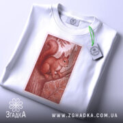 Футболка білка лісова марка, біла, зображена на столі, фон світлий, принт з білкою на дереві в червонувато-коричневих тонах, з етикеткою зеленого кольору.