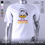 Футболка більше спати, біла, зображена з фронтального ракурсу на манекені, фон нейтральний, принт з мультяшним персонажем, що має сонний вираз обличчя, текст "ТРЕБА БІЛЬШЕ СПАТИ" червоного і жовтого кольорів.