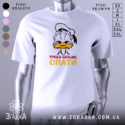 Футболка більше спати, біла, зображена з фронтального ракурсу на манекені, фон нейтральний, принт з мультяшним персонажем, що має сонний вираз обличчя, текст "ТРЕБА БІЛЬШЕ СПАТИ" червоного і жовтого кольорів.