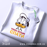 Футболка більше спати, біла, зображена зверху, на ній прикріплений етикет, фон світлий, принт з мультяшним персонажем, що має сонний вираз обличчя, текст "ТРЕБА БІЛЬШЕ СПАТИ" червоного і жовтого кольорів.