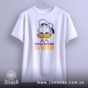 Футболка більше спати, біла, зображена з фронтального ракурсу, підвішена на вішалці, фон світлий, принт з мультяшним персонажем, що має сонний вираз обличчя, текст "ТРЕБА БІЛЬШЕ СПАТИ" червоного і жовтого кольорів.