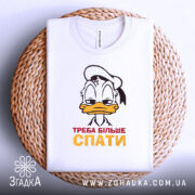 Футболка більше спати, біла, зображена зверху на плетеній кошику, фон світлий, принт з мультяшним персонажем, що має сонний вираз обличчя, текст "ТРЕБА БІЛЬШЕ СПАТИ" червоного і жовтого кольорів.