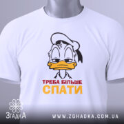 Футболка більше спати, біла, зображена зверху, на ній прикріплений етикет, фон світлий, принт з мультяшним персонажем, що має сонний вираз обличчя, текст "ТРЕБА БІЛЬШЕ СПАТИ" червоного і жовтого кольорів.