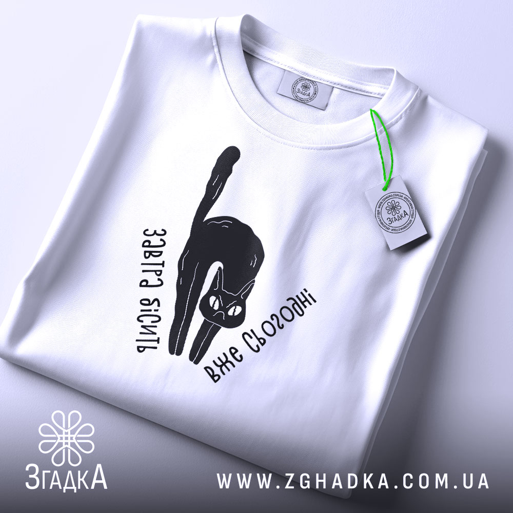 Футболка бісить вже сьогодні, складена біла футболка з чорним принтом кота, текстом 'Завтра бісить вже сьогодні', на фоні світло-сірого кольору, з зеленим ярликом.