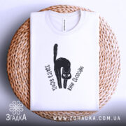 Футболка бісить вже сьогодні, складена біла футболка з чорним принтом кота, текстом 'Завтра бісить вже сьогодні', на плетеній основі, фон світло-сірий.