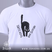 Футболка бісить вже сьогодні, складена біла футболка з чорним принтом кота, текстом 'Завтра бісить вже сьогодні', на фоні світло-сірого кольору, з зеленим ярликом.