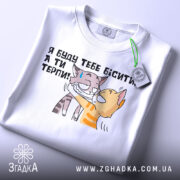 Футболка бісити терпіти, біла, складена на поверхні, фон світлий, принт з двома котами, один з котів з жовтими смужками, інший сірий, текст українською: 'Я буду тебе бісити, а ти терпи!', з етикеткою зеленого кольору.