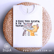 Футболка бісити терпіти, біла, складена на плетеній кошику, фон світлий, принт з двома котами, один з котів з жовтими смужками, інший сірий, текст українською: 'Я буду тебе бісити, а ти терпи!'