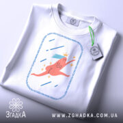 Футболка біжу волосся позаду, біла, складена на поверхні, фон світлий, принт з рожевим персонажем, що біжить, з короною та блакитним волоссям, прикріплена етикетка з логотипом.