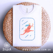 Футболка біжу волосся позаду, біла, складена на кошику з плетеного матеріалу, фон світлий, принт з рожевим персонажем, що біжить, з короною та блакитним волоссям, оточений зірочками та лініями.