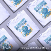 Футболка Blueberrinni Octopussini білого кольору, зображена з фронтального ракурсу на манекені, фон світло-сірий. Принт з веселим восьминогом, оточеним рослинними елементами та квітами, з короною на голові.