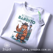 Футболка Bobritto Bandito білого кольору, складена на поверхні, фон світло-сірий, принт з ведмедем у капелюсі та пальто, що тримає гвинтівку, з етикеткою на зеленому шнурку.