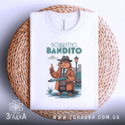 Футболка Bobritto Bandito білого кольору, складена на плетеній кошику, фон світло-сірий, принт з ведмедем у капелюсі та пальто, що тримає гвинтівку, на фоні вуличного ліхтаря.