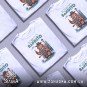 Футболка Bobritto Bandito білого кольору, зображена на манекені з правого боку, фон світло-сірий, принт з ведмедем у капелюсі та пальто, що тримає гвинтівку, на фоні вуличного ліхтаря та будинків.