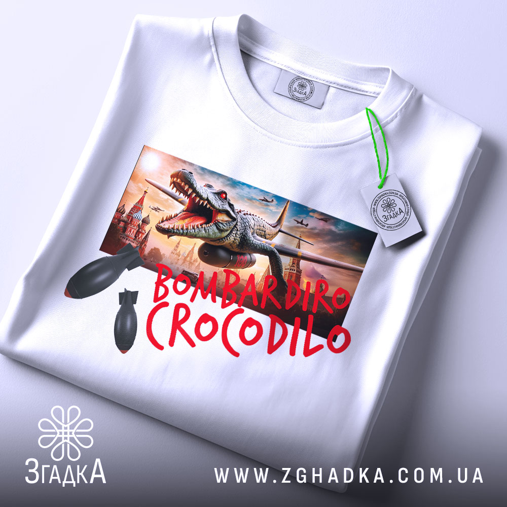 Футболка Bombardiro Crocodilo білого кольору, складена на поверхні, з тегом, принт з крокодилом та бомбами, фон світлий, деталі принта чіткі.