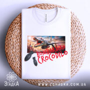 Футболка Bombardiro Crocodilo білого кольору, складена на плетеній кошику, фон нейтральний, принт з крокодилом та бомбами, деталі принта добре видимі.