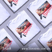 Футболка Bombardiro Crocodilo білого кольору, зображена на манекені з правого боку, фон нейтральний, принт з крокодилом, що тримає бомби, на фоні міського пейзажу.