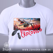 Футболка Bombardiro Crocodilo білого кольору, складена на поверхні, з тегом, принт з крокодилом та бомбами, фон світлий, деталі принта чіткі.