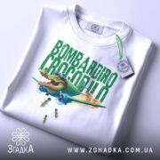 Футболка Bombardiro Crocodilo білого кольору, складена на столі, фон світло-сірий, принт з крокодилом в пілотських окулярах, що сидить в літаку, з зеленим текстом 'BOMBARDIRO CROCODILO' та бомбами, що падають, з етикеткою на шнурку.