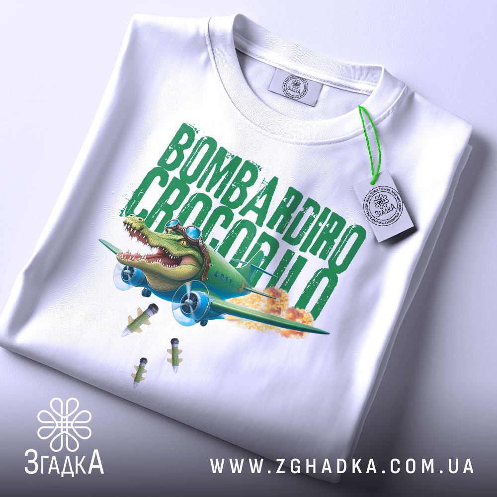 Футболка Bombardiro Crocodilo білого кольору, складена на столі, фон світло-сірий, принт з крокодилом в пілотських окулярах, що сидить в літаку, з зеленим текстом 'BOMBARDIRO CROCODILO' та бомбами, що падають, з етикеткою на шнурку.