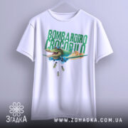 Футболка Bombardiro Crocodilo білого кольору, підвішена на вішалці, фон світло-сірий, принт з крокодилом в пілотських окулярах, що сидить в літаку, з зеленим текстом 'BOMBARDIRO CROCODILO' та деталями бомб, що падають.