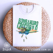Футболка Bombardiro Crocodilo білого кольору, складена на плетеному кошику, фон світло-сірий, принт з крокодилом в пілотських окулярах, що сидить в літаку, з зеленим текстом 'BOMBARDIRO CROCODILO' та бомбами, що падають.