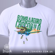 Футболка Bombardiro Crocodilo білого кольору, складена на столі, фон світло-сірий, принт з крокодилом в пілотських окулярах, що сидить в літаку, з зеленим текстом 'BOMBARDIRO CROCODILO' та бомбами, що падають, з етикеткою на шнурку.