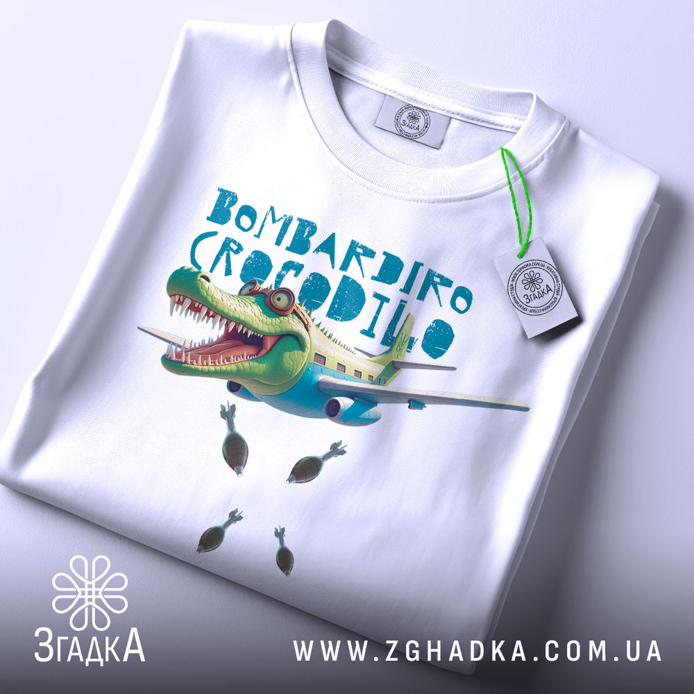 Футболка Бомбардіро Крокоділо білого кольору, складена на поверхні, зображена зверху, фон світлий, принт з крокодилом на літаку, з бомбами, на комірці є етикетка.