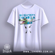 Футболка Бомбардіро Крокоділо білого кольору, підвішена на вішалці, фон світло-сірий, принт з крокодилом, що літає на літаку, з бомбами під ним.