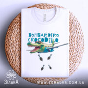 Футболка Бомбардіро Крокоділо білого кольору, складена на плетеній кошику, фон світлий, принт з крокодилом на літаку, з бомбами, вид з верхньої частини.
