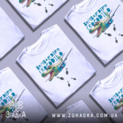 Футболка Бомбардіро Крокоділо білого кольору, зображена з фронтального ракурсу на манекені, фон нейтральний, принт з крокодилом, що літає на літаку, з бомбами під ним.