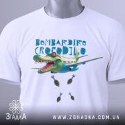 Футболка Бомбардіро Крокоділо білого кольору, складена на поверхні, зображена зверху, фон світлий, принт з крокодилом на літаку, з бомбами, на комірці є етикетка.