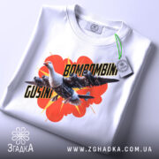 Футболка Bombombini Gusini білого кольору, складена на поверхні, з принтом птаха та літаків, на фоні червоного вибуху, з етикеткою, що звисає.