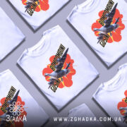 Футболка Bombombini Gusini білого кольору, зображена на манекені, з принтом, що містить птаха та літаки на яскравому фоні з червоними елементами.