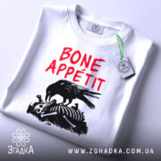 Футболка Bone Appétit білого кольору, складена на столі, ракурс зверху, фон світло-сірий, принт з чорним вороном, що клює кістки, текст червоним шрифтом 'BONE APPÉTIT', на ярлику зелена стрічка.