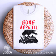 Футболка Bone Appétit білого кольору, складена на плетеному кошику, ракурс зверху, фон світло-сірий, принт з чорним вороном, що клює кістки, текст червоним шрифтом 'BONE APPÉTIT'.