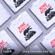 Футболка Bone Appétit білого кольору, зображена на манекені, ракурс спереду, фон світло-сірий, принт з чорним вороном, що клює кістки, текст червоним шрифтом 'BONE APPÉTIT'.