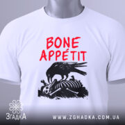 Футболка Bone Appétit білого кольору, складена на столі, ракурс зверху, фон світло-сірий, принт з чорним вороном, що клює кістки, текст червоним шрифтом 'BONE APPÉTIT', на ярлику зелена стрічка.