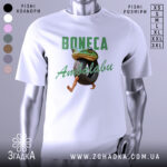 Футболка Boneca ambalabu білого кольору, зображена з фронтального ракурсу на манекені, фон світло-сірий, принт з жабою, що тримає шину, з написом 'Boneca Ambalabu' зеленого кольору.