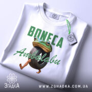 Футболка Boneca ambalabu білого кольору, зображена зверху на плоскій поверхні, фон світло-сірий, принт з жабою, що тримає шину, з написом 'Boneca Ambalabu' зеленого кольору, на футболці є етикетка з зеленим шнурком.