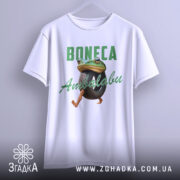 Футболка Boneca ambalabu білого кольору, зображена на вішалці, фон світло-сірий, принт з жабою, що тримає шину, з написом 'Boneca Ambalabu' зеленого кольору.