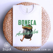 Футболка Boneca ambalabu білого кольору, зображена на плоскій поверхні, фон світло-сірий, принт з жабою, що тримає шину, з написом 'Boneca Ambalabu' зеленого кольору, футболка розташована на плетеній основі.