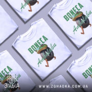 Футболка Boneca ambalabu білого кольору, зображена з фронтального ракурсу на манекені, фон світло-сірий, принт з жабою, що тримає шину, з написом 'Boneca Ambalabu' зеленого кольору.