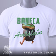 Футболка Boneca ambalabu білого кольору, зображена зверху на плоскій поверхні, фон світло-сірий, принт з жабою, що тримає шину, з написом 'Boneca Ambalabu' зеленого кольору, на футболці є етикетка з зеленим шнурком.