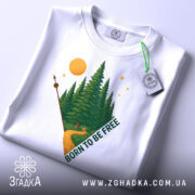 Футболка born free білого кольору, складена на поверхні, з принтом лісу, дороги та сонця, вид зверху, фон світло-сірий.