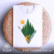 Футболка born free білого кольору, складена на плетеній кошику, з принтом лісу, дороги та сонця, вид зверху, фон світло-сірий.